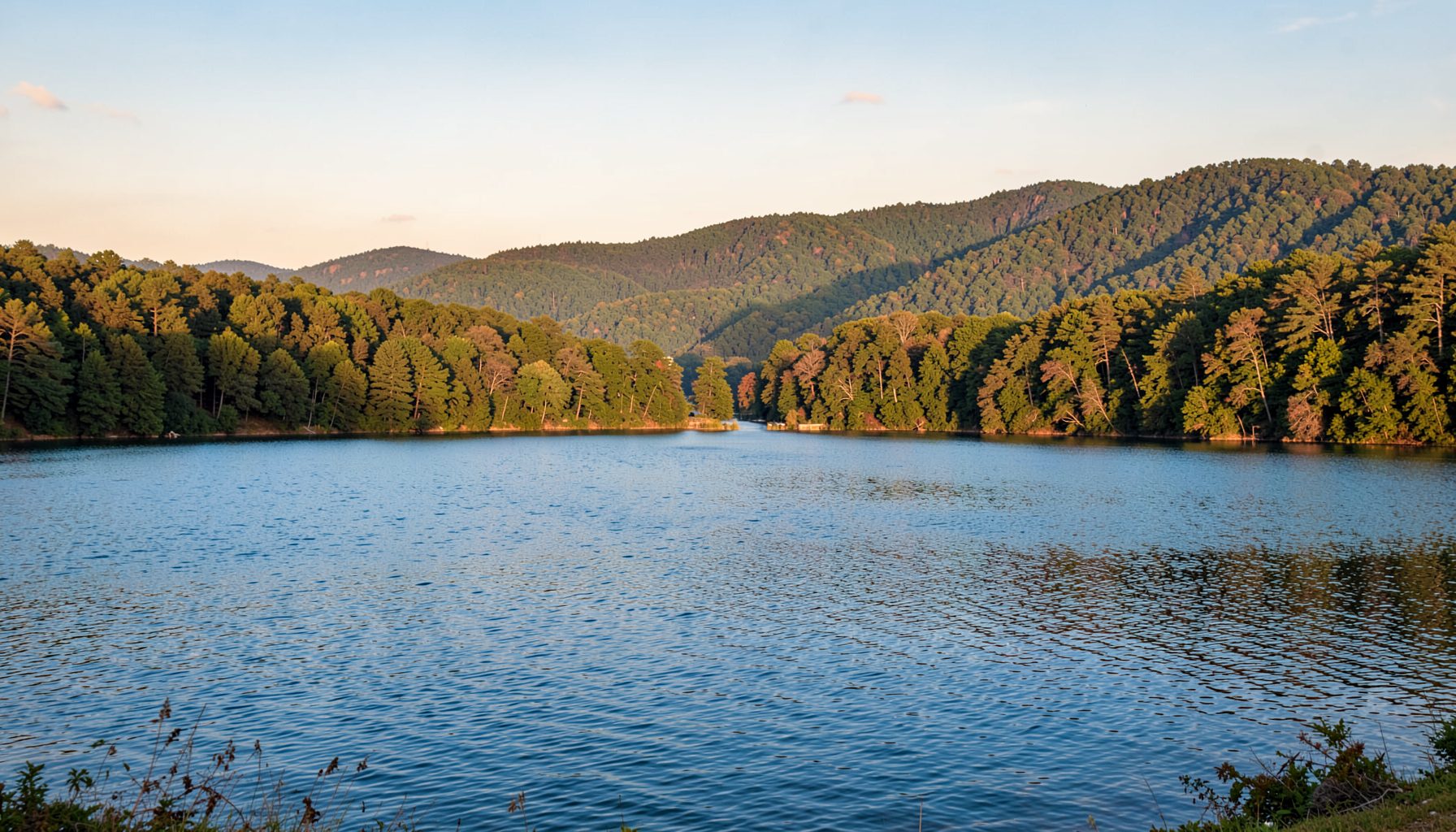 A sweeping landscape view of Caesar Creek State Park with calm blue lake water in the foreground, green wooded hills rolling into the distance under a soft sky, warm earthy tones throughout