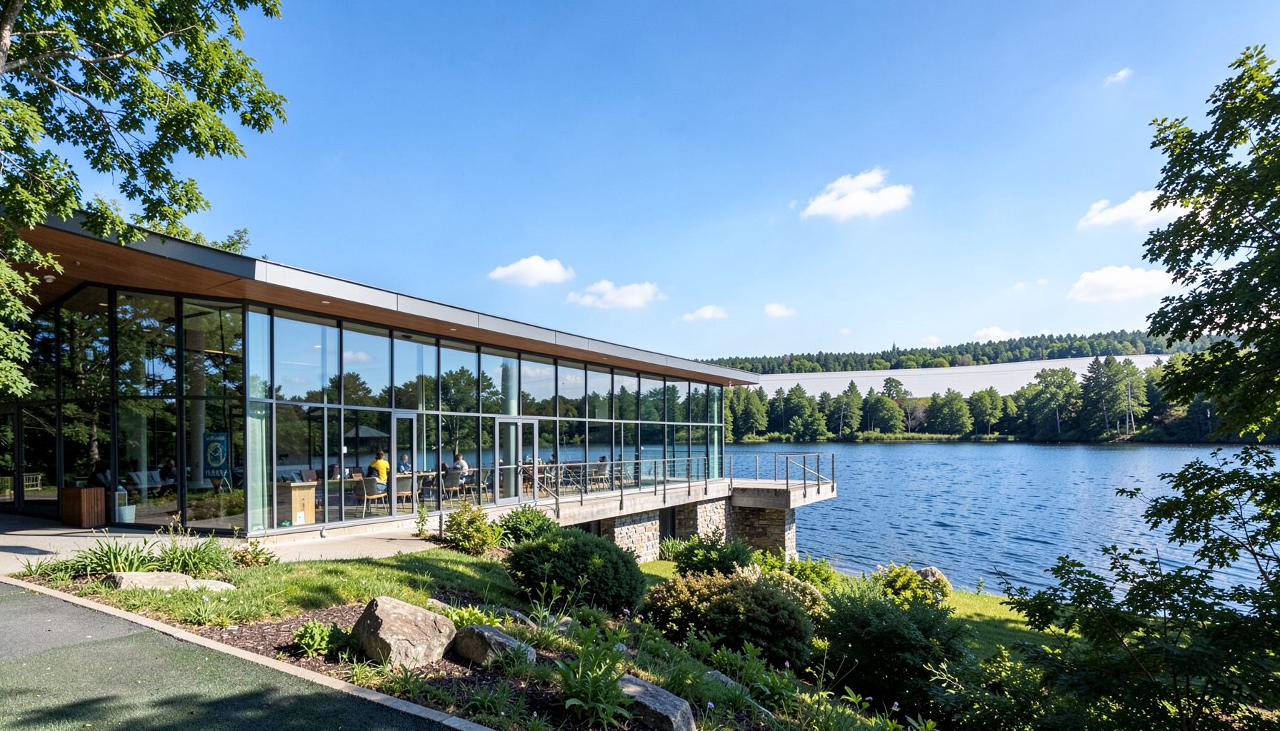 A modern visitor center building with large windows overlooking a lake, surrounded by trees under a blue sky